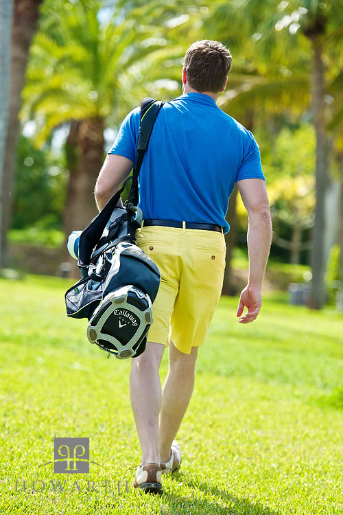 A golfer walks to the next tee carrying his golf bag dressed casually in bright yellow Bermuda shorts and a blue polo shirt