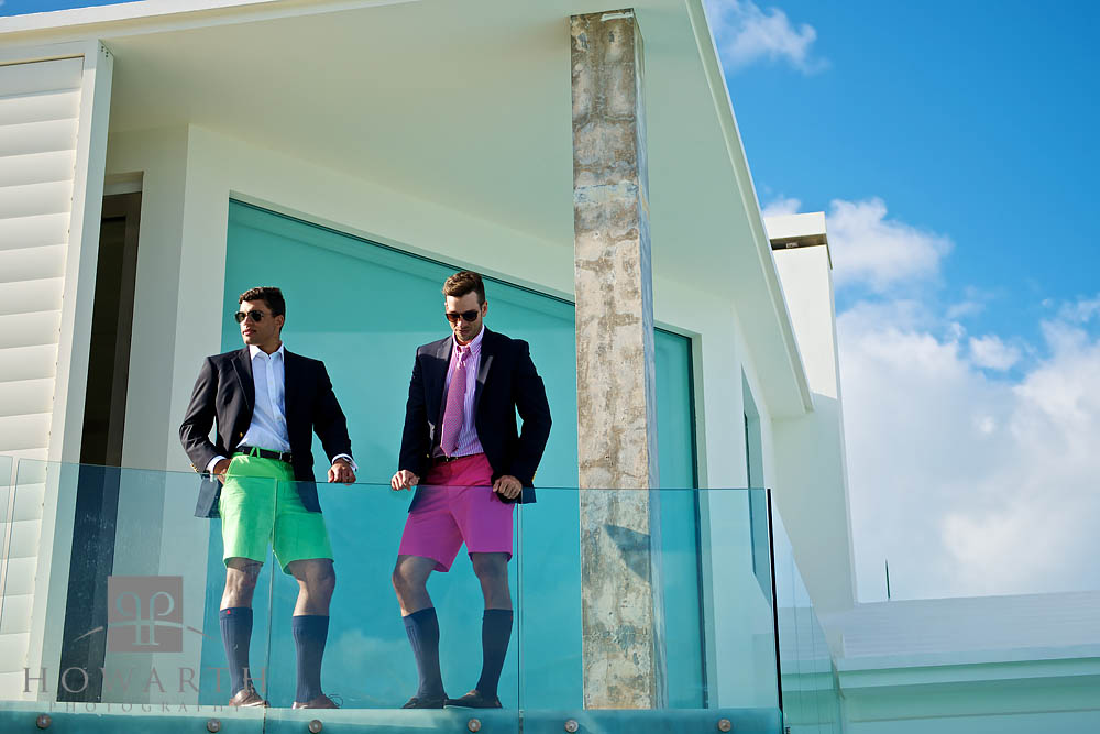 Two young professionals standing on a glass front balcony wearing brightly colored green and pink Bermuda Shorts, jackets and...